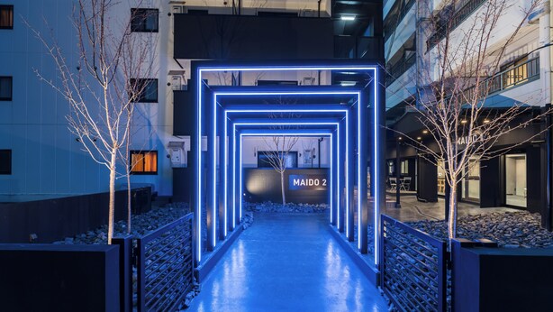 Una entrada de edificio moderno con un camino iluminado por neón que lleva a un letrero que dice 'MAIDO 2'.