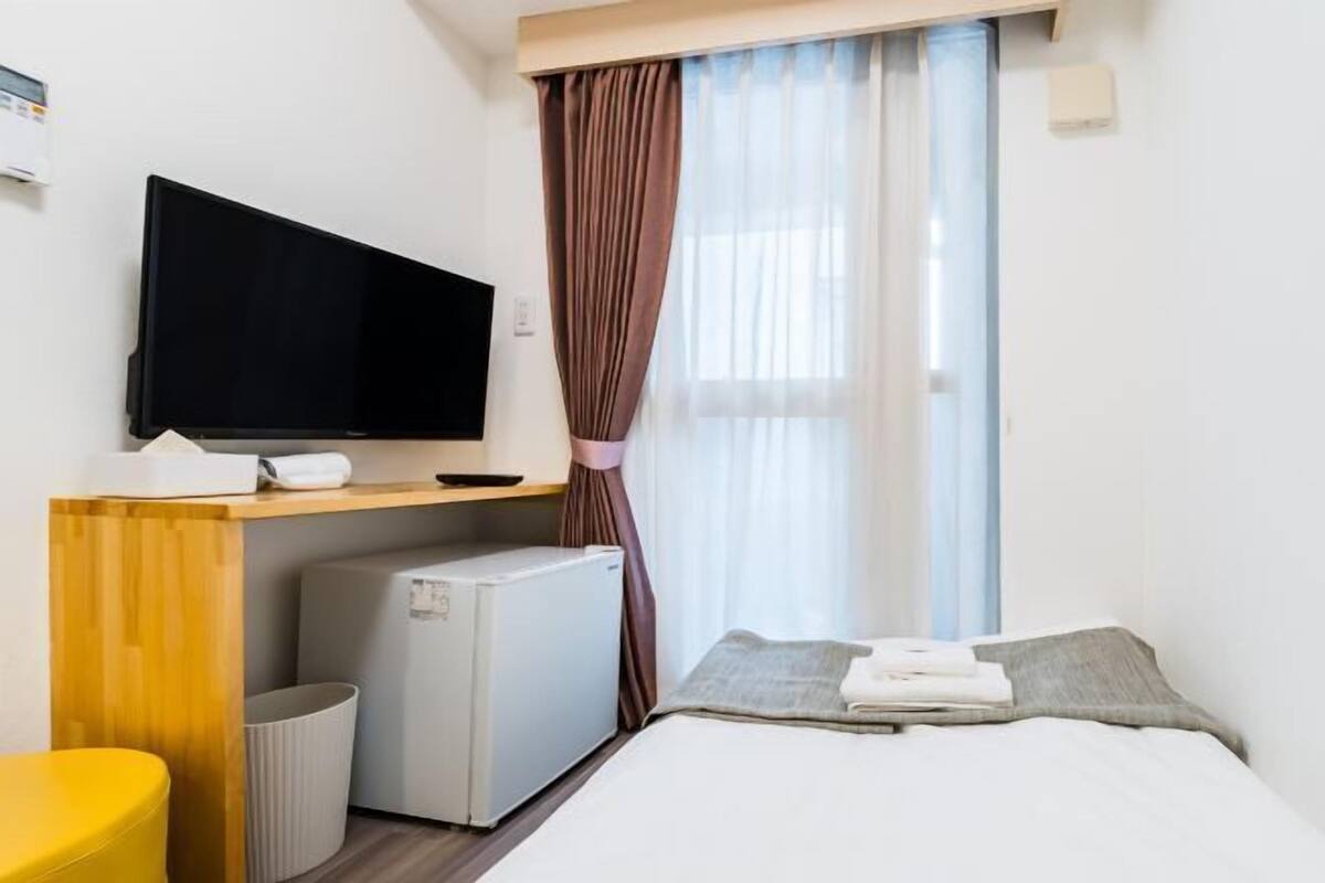 A compact hotel room with a bed, a small desk with a flat-screen TV, a mini-fridge, a trash bin, and a yellow chair.