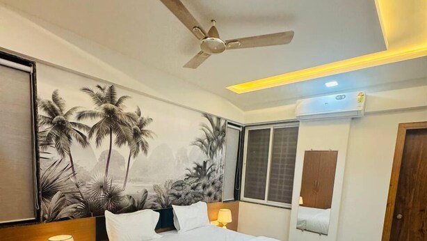 A hotel room with a bed, two bedside tables with lamps, a ceiling fan, a wall mural, an air conditioning unit, and a door. - Mighter Crest Studio Apartment (Nashik)