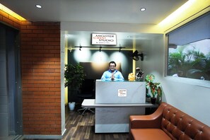 A reception desk with a sign for MIGHTER CREST STUDIO by BEEQTEL, a black office chair, a potted plant, and a window with a view of palm trees. - Mighter Crest Studio Apartment (Nashik)