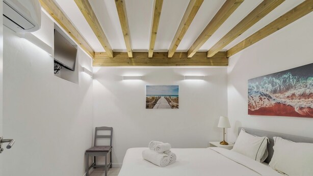 Une chambre d’hôtel avec un plafond en bois, un lit recouvert de draps blancs, une chaise, une table de chevet avec une lampe et un tableau accroché au mur.