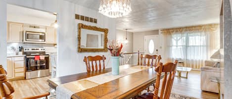 Uma sala de jantar espaçosa com mesa e cadeiras de madeira, um candelabro e vista para a cozinha ao fundo.