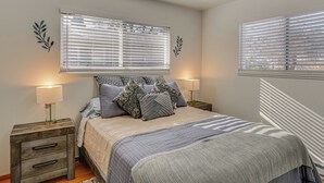 Un dormitorio con una cama, dos mesitas de noche con lámparas y dos ventanas con persianas.