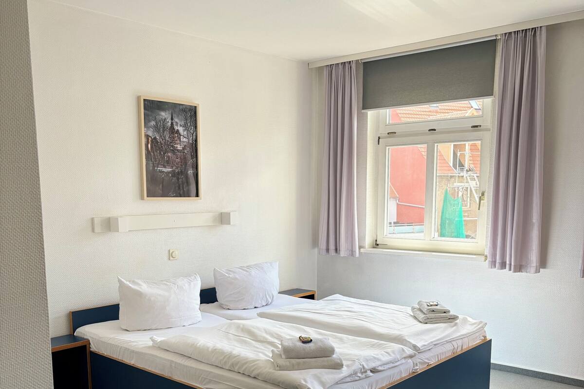 A neatly made bed with white linens and a folded towel, a bedside table, a framed picture on the wall, and a window with curtains.