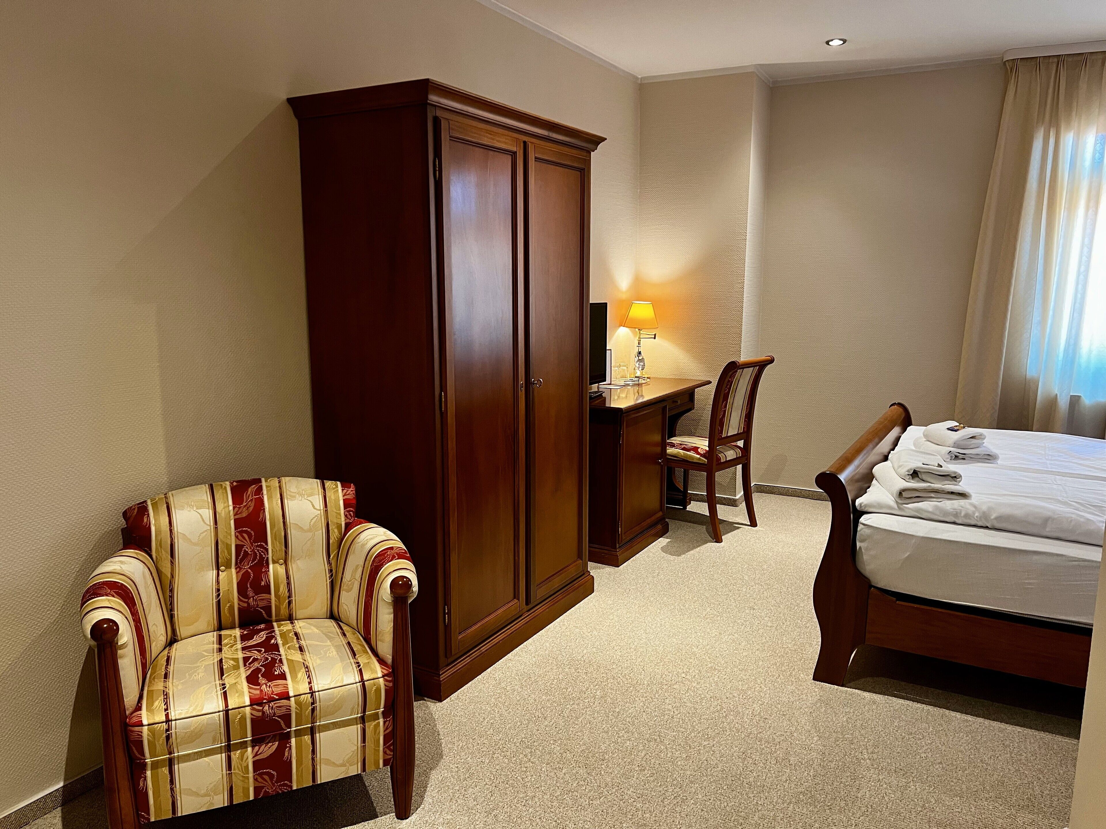 Una habitación de hotel con una cama, un escritorio con lámpara, un armario y un sillón a rayas.