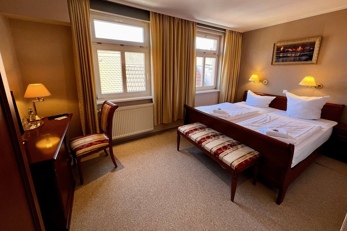 A hotel room with a bed, bedside lamps, a desk with a lamp, a chair, a bench, and a window with curtains.