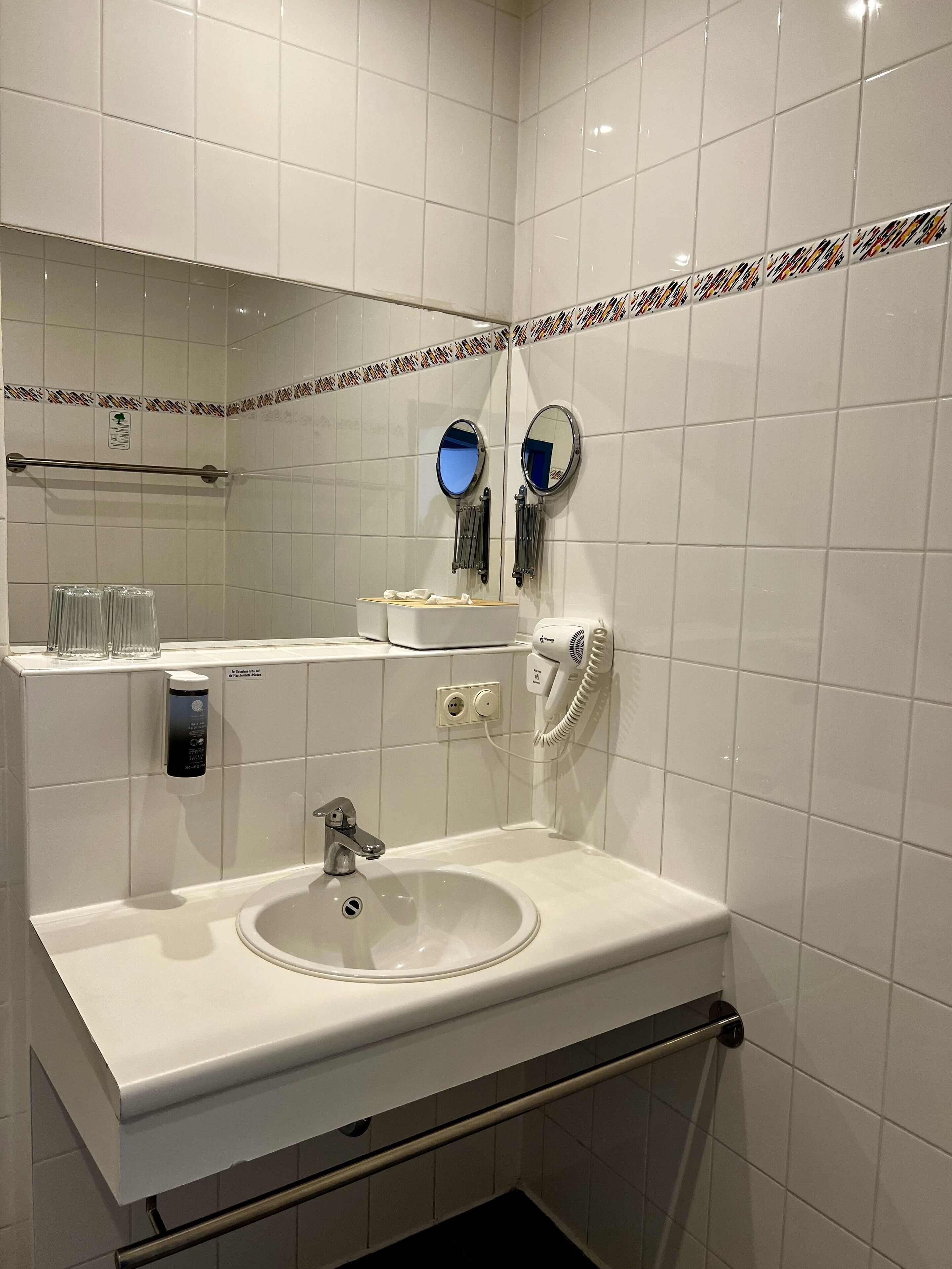 Une salle de bain moderne avec un lavabo blanc, deux miroirs, un distributeur de serviettes et un sèche-cheveux.