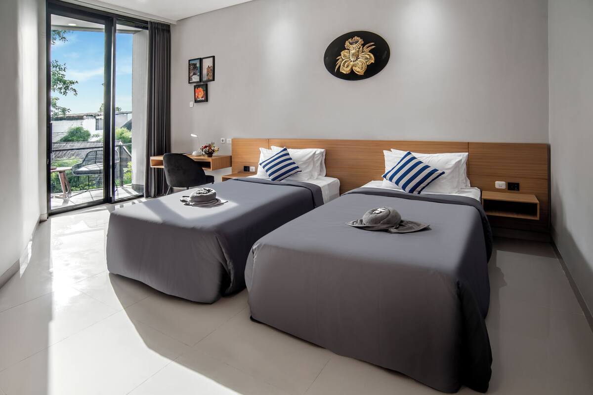 A modern hotel room with two beds, a desk, and a view of the outdoors.