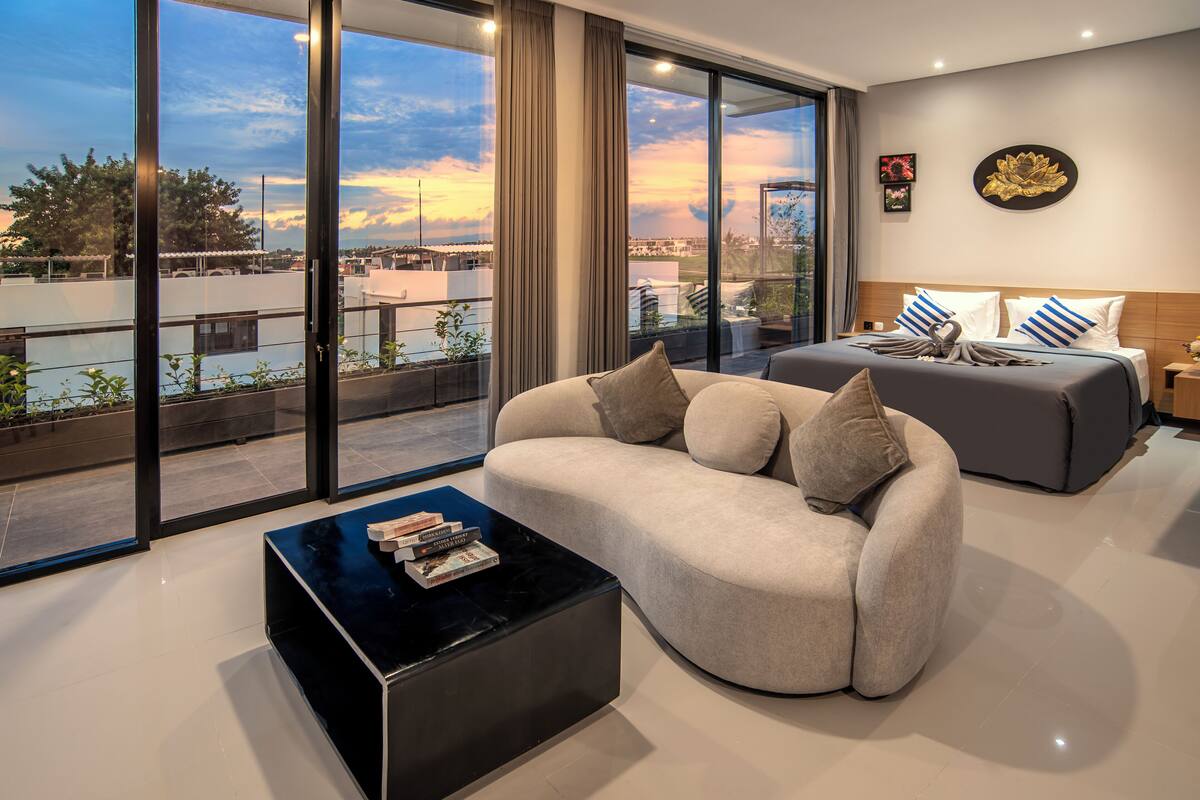 A modern hotel room with a large sliding glass door leading to a balcony, a sofa, a coffee table with books, and a bed with decorative pillows.