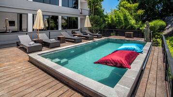 A modern outdoor swimming pool with lounge chairs and umbrellas, surrounded by a wooden deck and lush greenery.