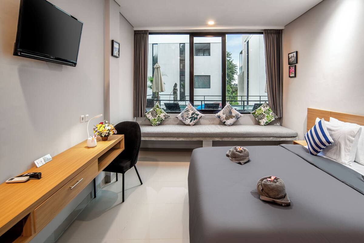A modern hotel room with a bed, a desk, a chair, a TV, and a window with a view of a building and a balcony.