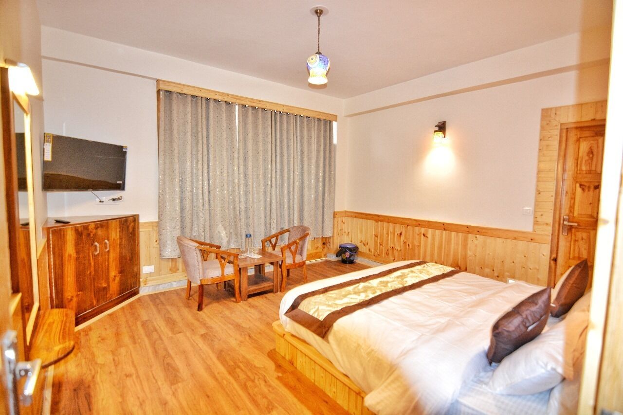 Une chambre d’hôtel avec un lit en bois, un bureau avec des chaises, une télévision et une fenêtre avec des rideaux.