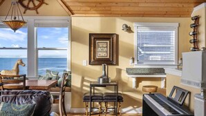 Un salón acogedor con vista al océano, un piano, una mesa con sillas y un techo de madera.