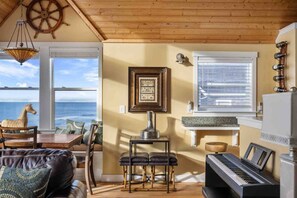 Uma sala de estar aconchegante com vista para o oceano, um piano, uma mesa com cadeiras e teto de madeira.
