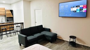 Un soggiorno moderno con un divano componibile, una TV a schermo piatto montata a parete, un angolo cottura con forno a microonde e un tavolo da pranzo con sgabelli.