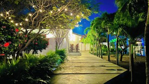 Un vialetto conduce a un edificio illuminato da luci a tenda, circondato da una vegetazione rigogliosa e palme.