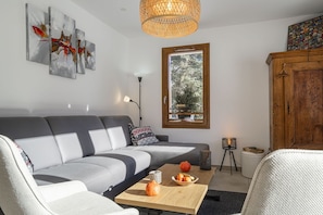 A modern living room with a grey sofa, a wooden coffee table with fruits, a lamp, and a window with a view of trees.