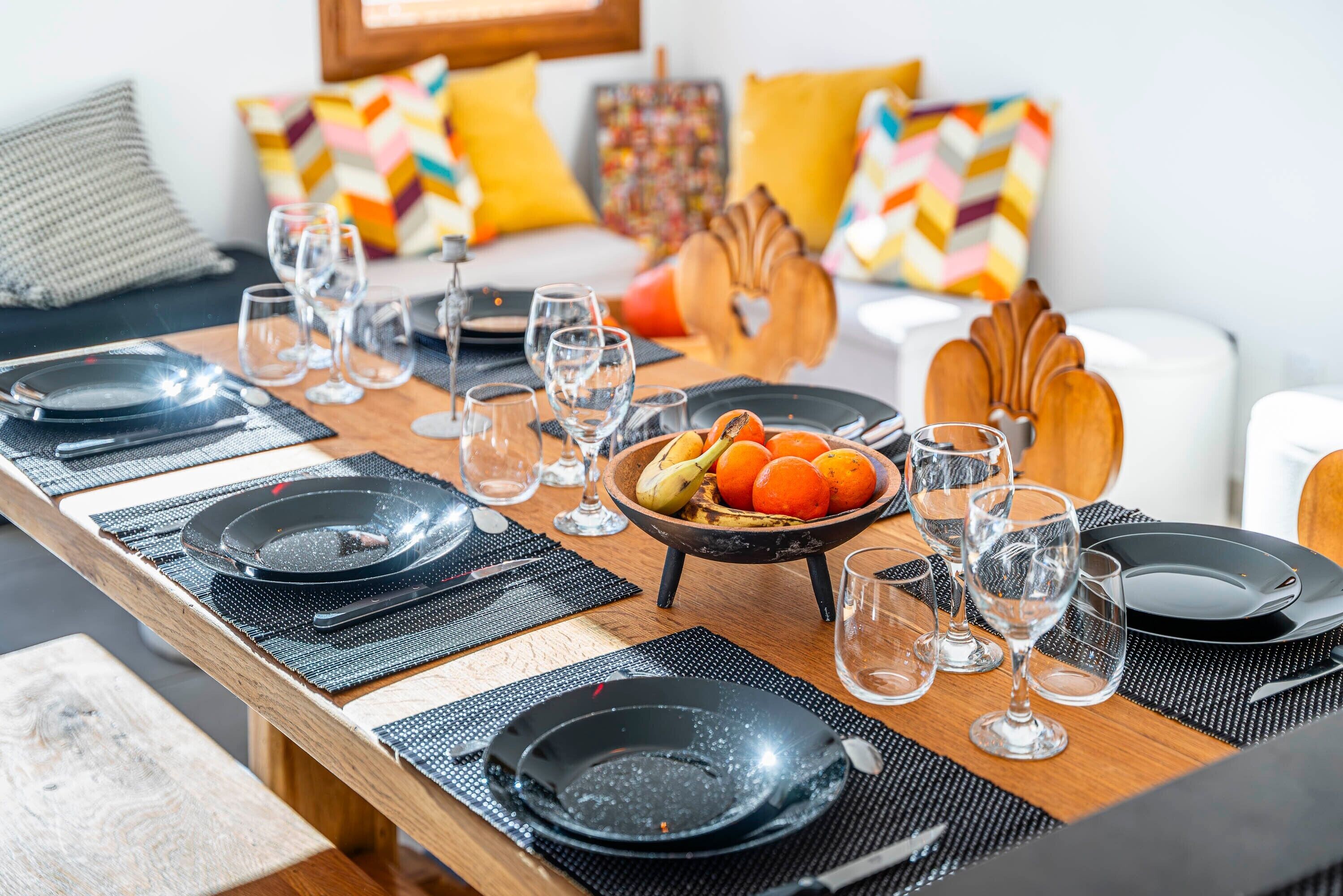 Une table à manger dressée avec des assiettes noires, des verres transparents et un saladier rempli de fruits.