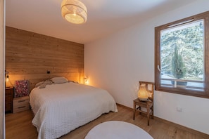 A bedroom with a wooden headboard, a bed with a white blanket, a bedside table with a lamp, and a window with a view of trees.