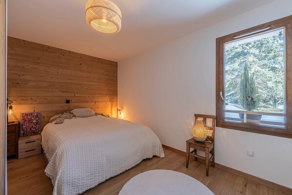 A bedroom with a wooden headboard, a bed with a white blanket, a bedside table with a lamp, and a window with a view of trees.