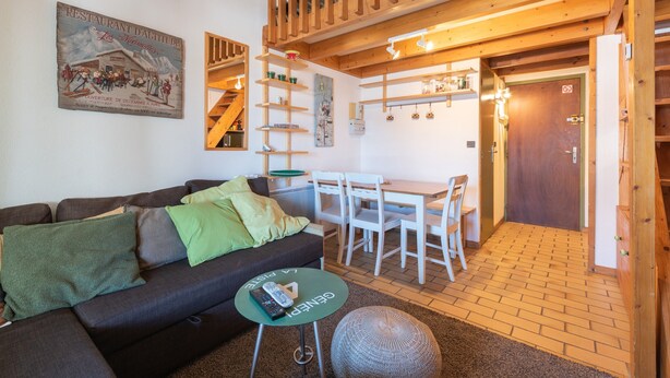 A cozy living room with a sofa, a small round table, a dining area with chairs, and a staircase leading to an upper level.
