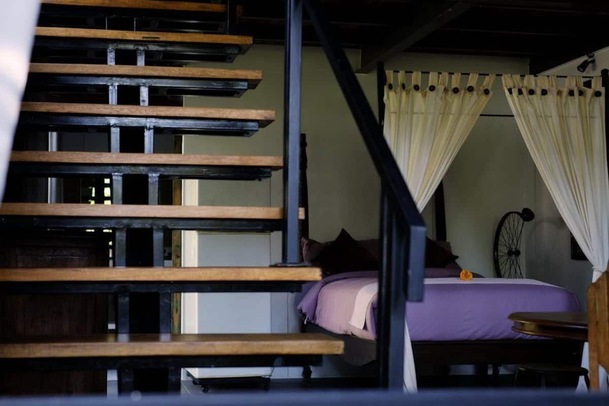 A bedroom with a bed, a nightstand, a chair, a window with curtains, and a staircase with wooden steps.