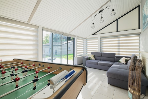 A modern living room with a foosball table, a gray sofa, and large windows.
