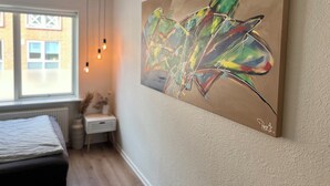 A room with a bed, a small bedside table, a nightstand, and a window with a view of buildings. - Cozy Flats Silkeborg - S1 (Silkeborg)