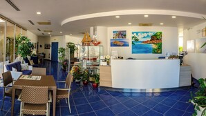 A hotel reception area with a curved reception desk, blue-tiled floor, and various potted plants. - Astro Suite (Cefalù)