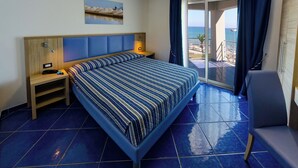 A hotel room with a large bed, a nightstand, a desk, and a chair. There is a view of the beach and the ocean through the balcony doors. - Astro Suite (Cefalù)