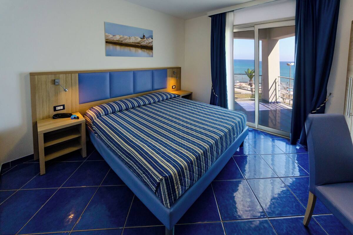 A hotel room with a large bed, a nightstand, a desk, and a chair. There is a view of the beach and the ocean through the balcony doors.