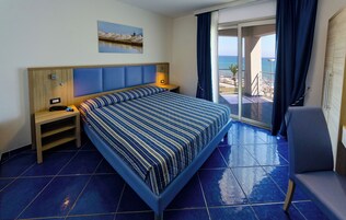 Habitación de hotel con una cama grande, una mesita de noche, un escritorio y una silla. Se ve la playa y el océano a través de las puertas del balcón.