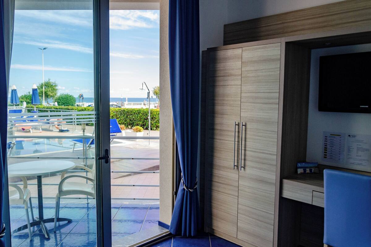 A room with a view of a pool and cityscape, a wooden wardrobe, a desk with a flat-screen TV, and a glass door.