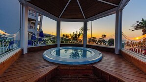 A wooden deck with a hot tub, glass railings, and a view of a sunset over a beach. - Astro Suite (Cefalù)