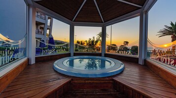 Una terraza de madera con un jacuzzi, barandas de vidrio y vistas a una puesta de sol sobre la playa.