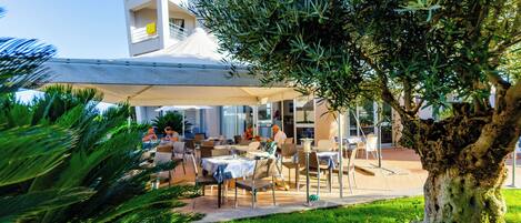 An outdoor seating area with tables and chairs under a canopy, surrounded by greenery and a building with balconies.