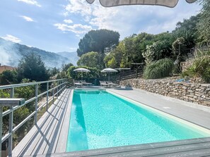 Ein rechteckiger Pool mit Holzterrasse und Metallgeländern, umgeben von üppiger Vegetation und einer Steinmauer.