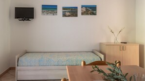 Une petite pièce avec un lit, un téléviseur fixé au mur et trois tableaux représentant des plages.