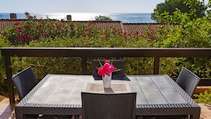 En terrasse med et bord og stole, en vase med blomster, og udsigt til havet.