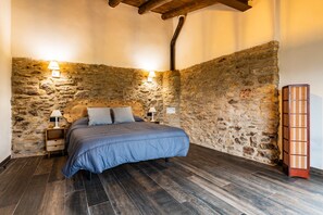 Een slaapkamer met een stenen muur, een bed met een blauw beddegoed, twee bedlampen en een houten nachtkastje.
