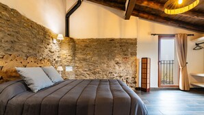 Un dormitorio con pared de piedra, una cama con cubrecama oscuro, una mesita de noche, una lámpara y vistas al exterior.