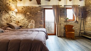 Ein Schlafzimmer mit Steinwand, Holzbalken, einem Bett, einem Sessel und einem Fenster mit Vorhängen.