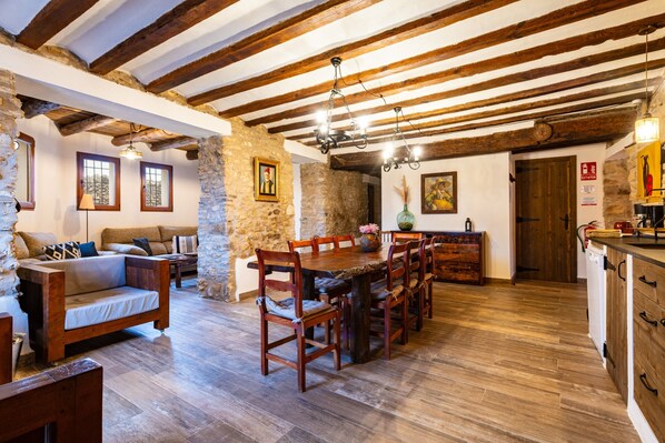 A rustic interior with exposed wooden beams, a stone wall, a dining table with chairs, a sofa, and a kitchen area.