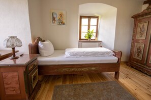 Una cama de madera con ropa de cama blanca, una lámpara de noche, una ventana con una planta y un mueble de madera con cuadros enmarcados.