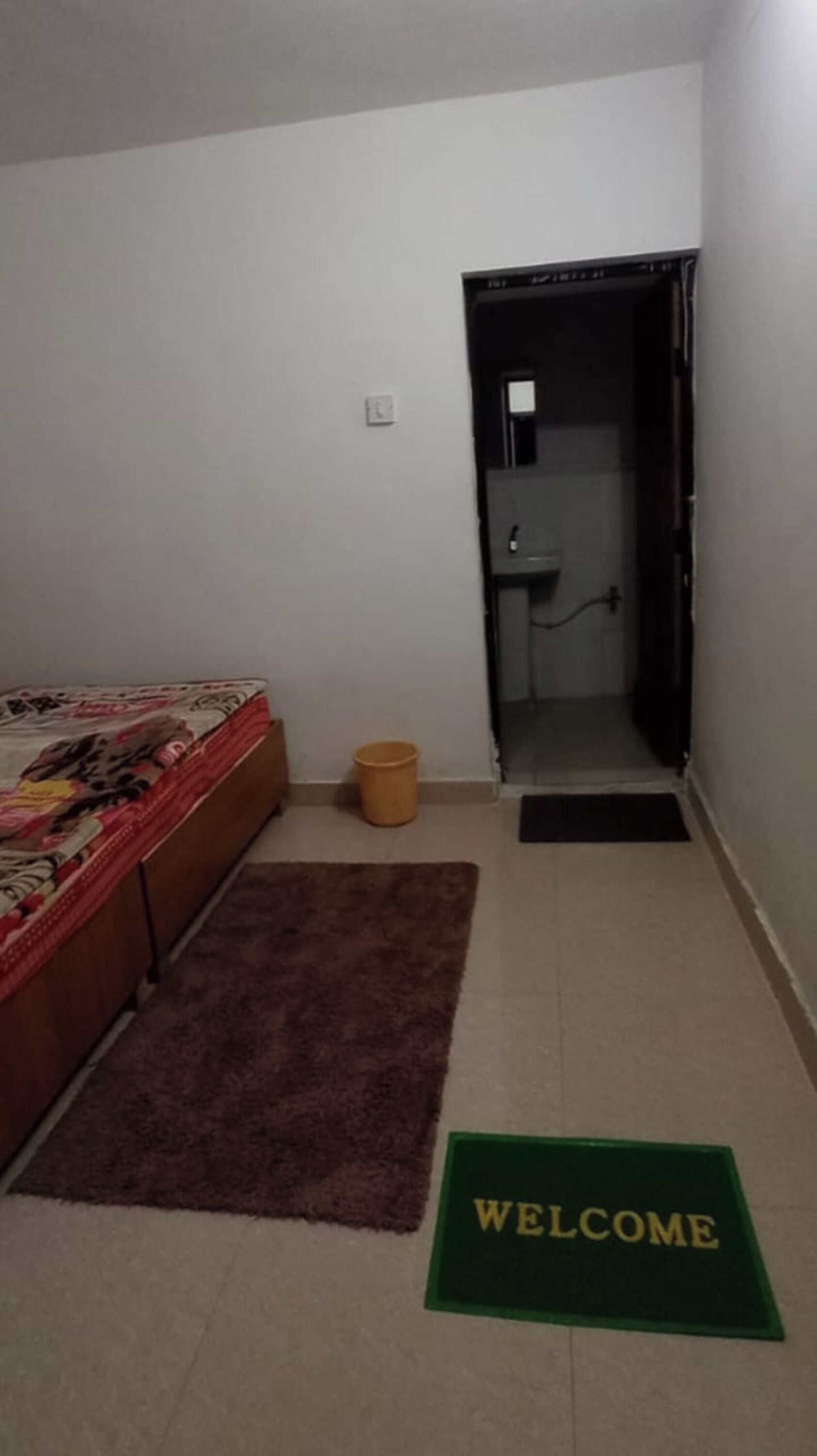 A small, dimly lit room with a bed, a welcome mat, and a door leading to a bathroom.