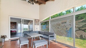 Une terrasse avec un canapé gris, une table blanche et un banc, une carte accrochée au mur et une vue sur l’extérieur.