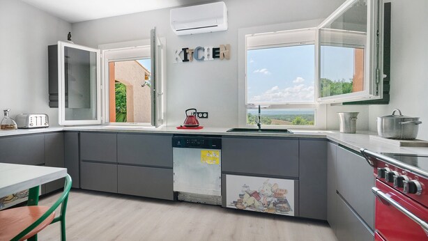 Une cuisine moderne avec des meubles gris, un fourneau rouge, un lave-vaisselle et une fenêtre donnant sur un beau paysage.