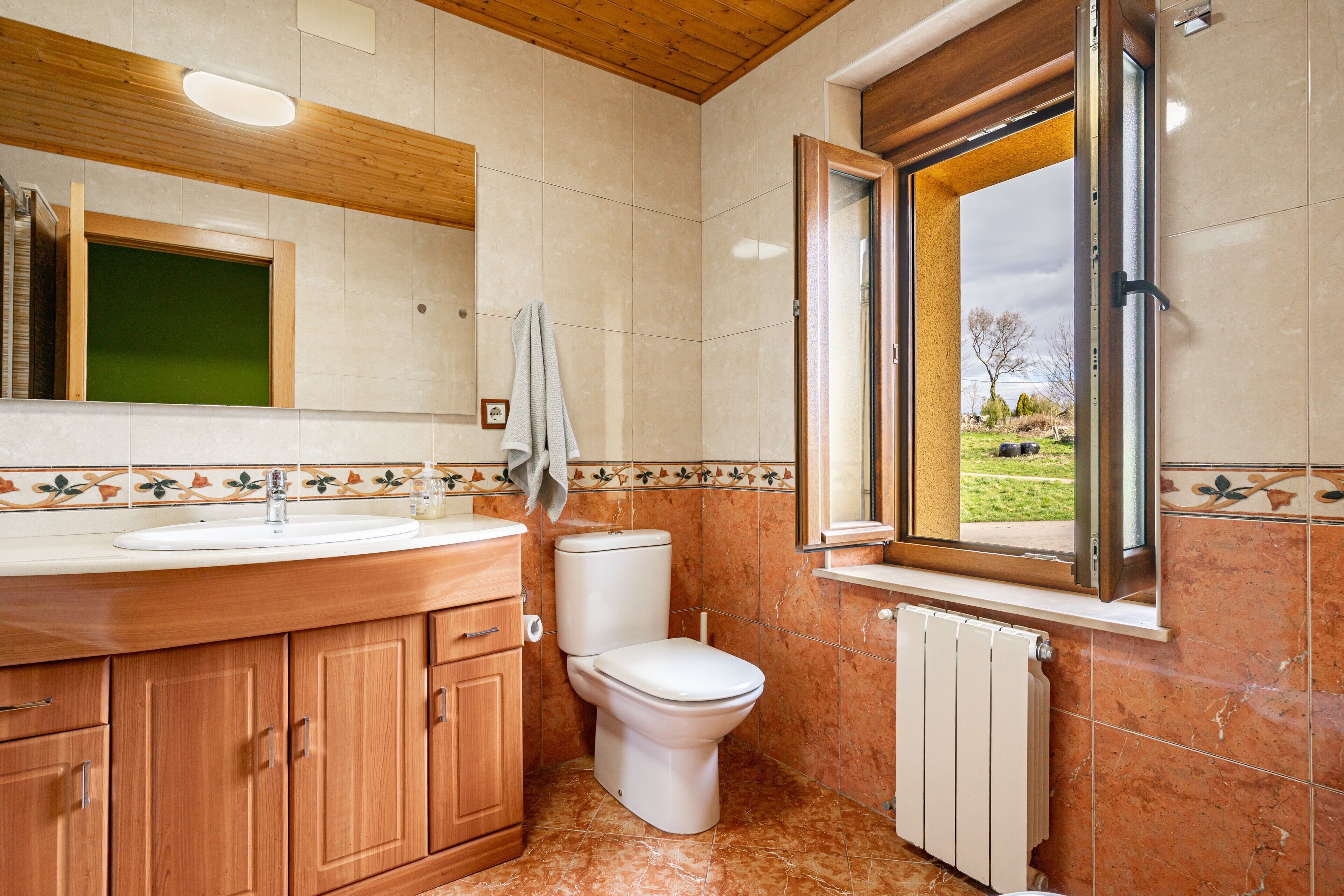 Een badkamer met een houten wastafelmeubel, een toilet, een raam met uitzicht en een betegelde spatwand.