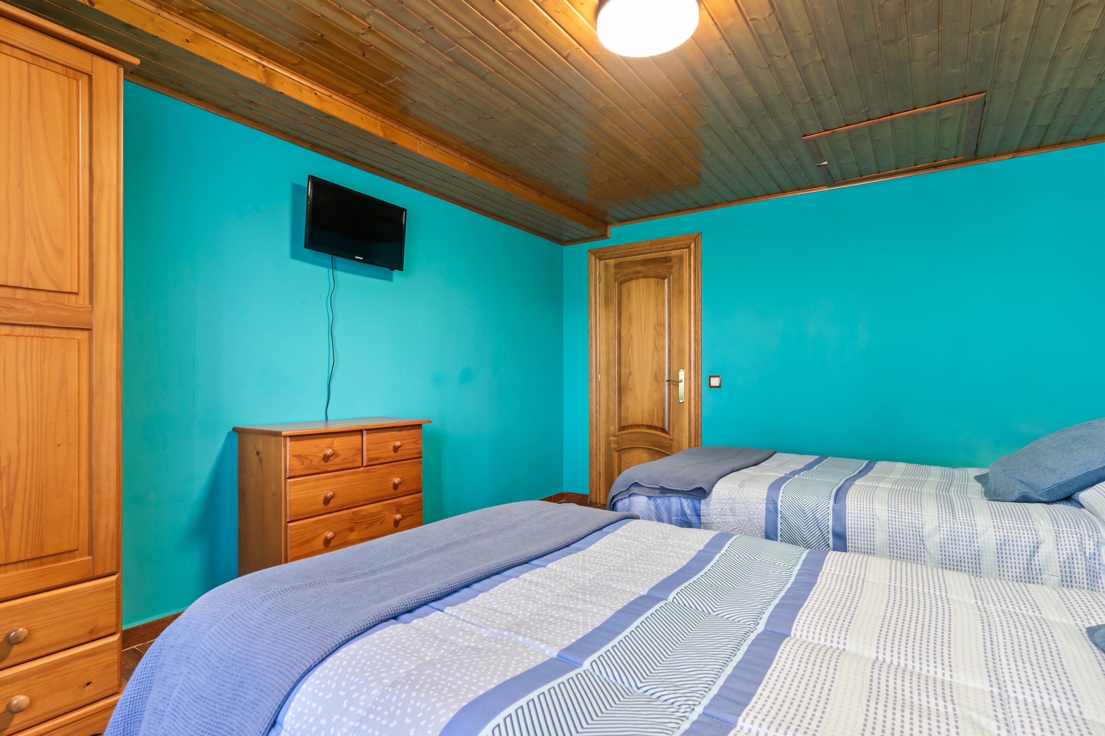 A room with teal walls, wooden ceiling, a TV mounted on the wall, a wooden dresser, and two beds with blue bedding.