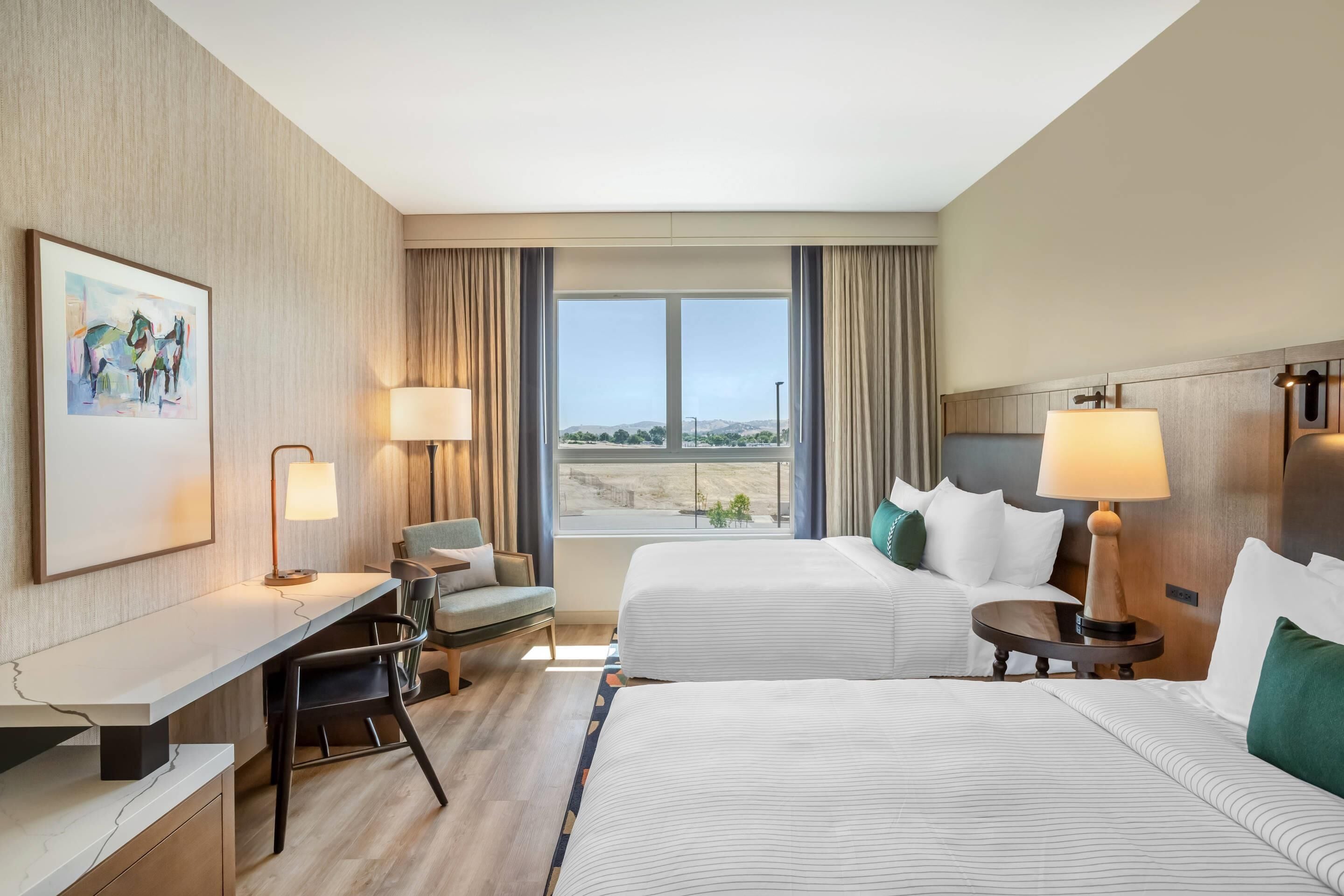 A hotel room with two beds, a desk, a chair, a lamp, and a large window with a view of the landscape.